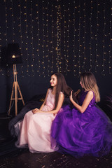 Cheerful girlfriends. Two girls comb each other's hair, sitting side by side in studio on dark...