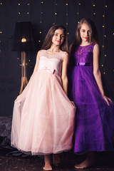 Cheerful girlfriends. Two long-haired girls in pink and purple dresses in studio on dark background...