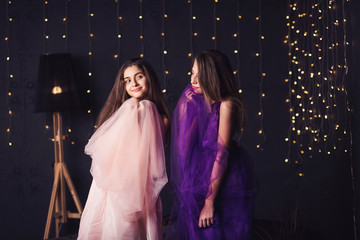 Cheerful girlfriends. Two long-haired girls in pink and purple dresses in studio on dark background with bokeh. Copy space.