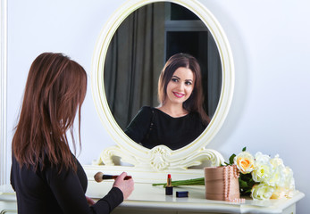 Portrait of beautiful young smiling brunette woman, wearing black dress, applying make-up, sitting...