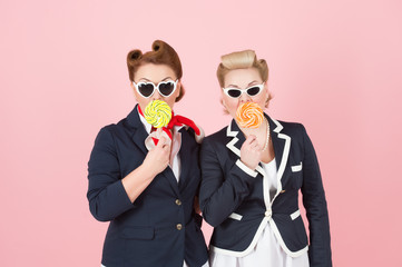Cheerful girls couple with lollipops and glasses. Girls hide mouth by lollipops and eyes by glasses. Official women in non official style. Pinup styled women with curls in blue jackets and sunglasses