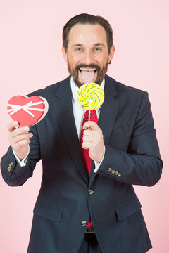 businessman with lollipop and heart in hands. portrait of handsome man holding candy and opened mouth with tongue. young bearded man going to eat lollipop. guy in blue suit holds present box on pink