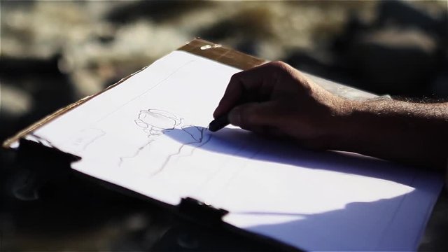 Close-Up of a Caucasian Male Hand Drawing and Erasing with a Black Pencil on White Paper. Filmed Outside. 