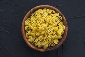 Macaroni ruote pasta in a wooden bowl on a black textured background, in the center close-up from the top.