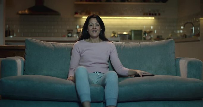 Young, attractive woman sits down on a sofa and turns on the television using a remote control.