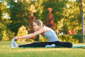 young pretty girl doing stretching on the park's grass. Wearing sport clothes in vlack and white colors. green summer or spring park on background. Feeling relaxed, chilled, happy.