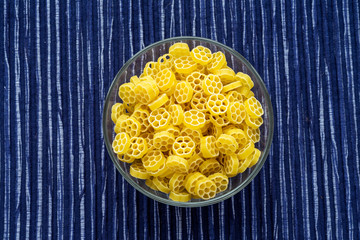 Macaroni ruote pasta in a glass bowl on a striped white blue fabric background in the center. Close-up with the top.