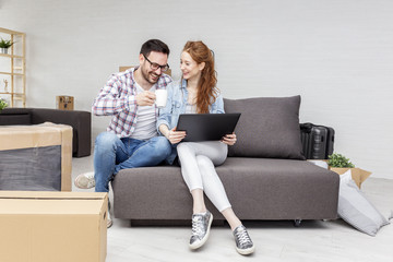 Young couple sitting in new empty room with unpacked boxes and looking at laptop while he drink cofe from mug