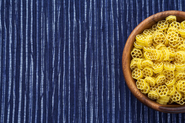 Macaroni ruote pasta in a wooden bowl on a striped white blue cloth background with a side. Close-up with the top. Free space for text.