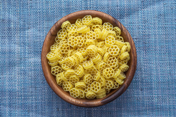 Macaroni ruote pasta in a wooden bowl on a blue knitted background in the center. Close-up with the top.