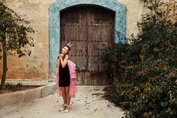Fototapeta na wymiar a sweet princess girl in a crown and a burgundy dress with a pink veil is pampered in front of an old house with an ancient wooden front door and a huge overgrown bush of dog rose