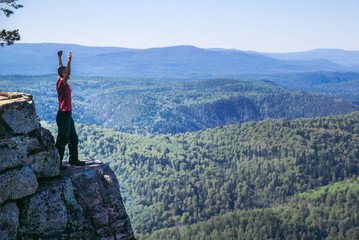 Young man hiker standing on top of a mountain with raised up hands, celebrating success. Full length shoot with copy space. Summer vacation and extreme activity