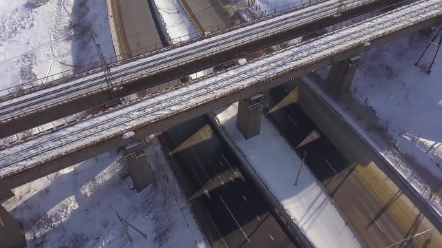 Suspension railway bridge above winter highway and car traffic on snowy road aerial landscape. Drone view train bridge and car moving on highway road.