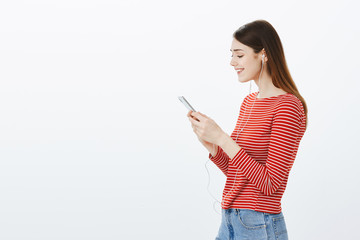 Fototapeta premium Girl recalling good memories, looking at photos in smartphone album. Portrait of pretty european female student, standing in profile, listening music in earphones or watching video in smartphone