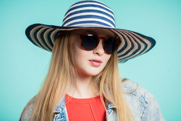 Young blonde woman with sunglasses and striped hat on blue background