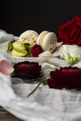 Three macaroons pastry lying on a plate surrounded by rose blossoms and rose petals on a wooden table covered with a crumpled white cloth