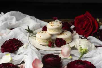 Three macaroons pastry lying on a plate surrounded by rose blossoms and rose petals on a wooden table covered with a crumpled white cloth
