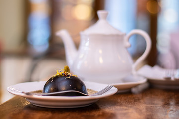Italian dessert: round chocolate cake with pistachio and dry petals of violets, served on a white plate, next to a white teapot on wooden table