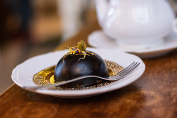 Italian dessert: round chocolate cake with pistachio and dry petals of violets, served on a white plate, next to a white teapot on wooden table