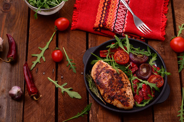 Grilled chicken breast with green salad from arugula and vegetables on a black pan. Wooden rustic background. Top view