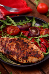 Grilled chicken breast with green salad from arugula and vegetables on a black pan. Wooden rustic background. Top view