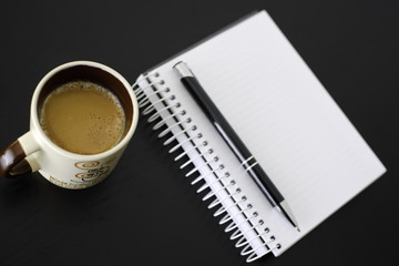 Coffee, notebook and pen on black wooden table. Coffee is an important drink in the workplace.