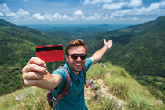 Young smiling male showing empty credit card. He is standing on top of mountain and showing the outdoor beauty.