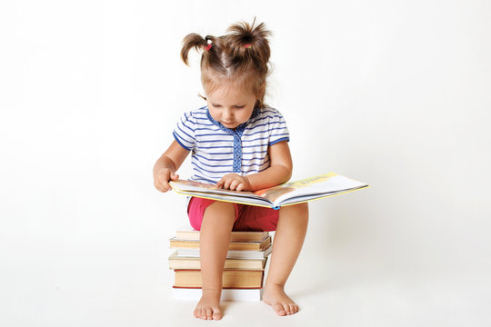 Curious female child with two funny pony tails, sits on pile of books, reads interesting faity tale, views colourful pictures with great interest, learns to read isolated over white studio background.