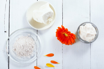 sea clay powder, moisturizer and raw shea butter block with gerbera flower on white wooden table background