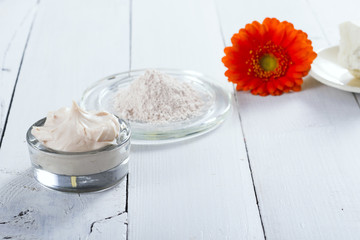 sea clay powder, moisturizer and raw shea butter block with gerbera flower on white wooden table background