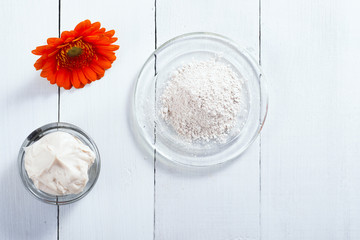sea clay powder, moisturizer and raw shea butter block with gerbera flower on white wooden table background