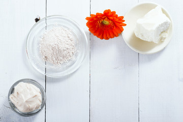 sea clay powder, moisturizer and raw shea butter block with gerbera flower on white wooden table background