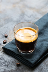 Glass cup of fresh coffee with thick golden foam froth on dark rustic background, espresso americano on black tissue with spoon of brown sugar and coffee beans, vertical copy space