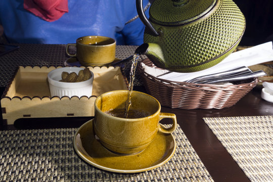 green tea from a old shabby cast iron kettle is poured into a cup closeup on a blurred background of the second full cup on the opposite side of the table