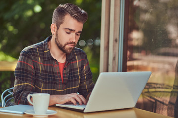 A handsome fashionable male freelancer with stylish haircut and beard, wearing fleece shirt, working on a laptop computer inside a cafe.