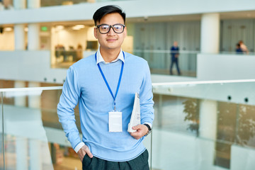Smiling Asian manager with hand in pocket looking at camera while standing at spacious office lobby, waist-up portrait shot