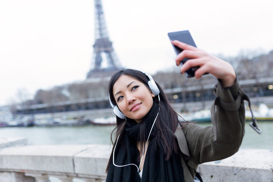 Pretty asian young woman taking a selfie in front of Eiffel tower in Paris while listening to music.