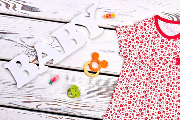 Infant girl clothes, accessories. Baby-girl small flowers print cotton dress, pacifier, teether, hair accessories, white wooden background.