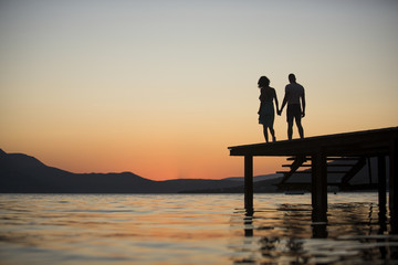 Fototapeta na wymiar Silhouette of sensual couple stand on pier with sunset above sea surface on background. Couple in love on romantic date in evening at dock, copy space. Romance and love concept.