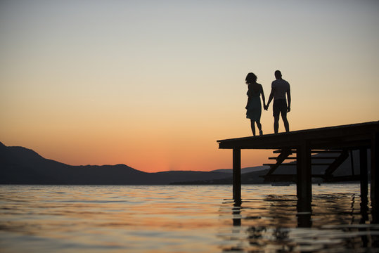 Silhouette of sensual couple stand on pier with sunset above sea surface on background. Couple in love on romantic date in evening at dock, copy space. Romance and love concept.