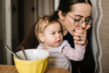 Young happy mother feeding her little baby with delicious tasty soup from plate in kitechen. Funny portrait of eating child. Beautiful girl in glasses together with daughter have diinner at home.