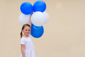 Fototapeta na wymiar Close-up of cute caucasian girl standing, smiling and playing with blue and white balloons. She wears white dress, and has braided hair. She is having fun.