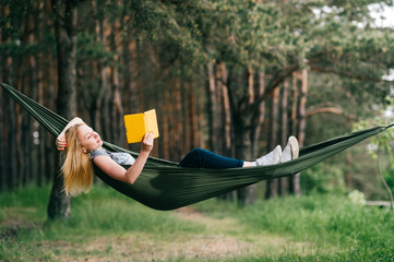 Young beautiful blonde girl in hammock reading e-book. Pretty woman leisure lifestyle portrait at...