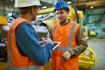 Two young technicians wearing reflective vests and hardhats using digital tablet while discussing how to solve problems with workflow, interior of spacious production department on background