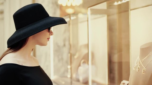 A beautiful young stylish lady in a hat and sunglasses approaches the shop window of a boutique and looks at goods with interest. Luxury jewelry store