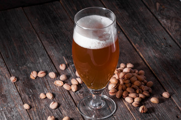 A glass of refreshing light beer and a scattered handful of pistachios on a table of old textured wooden boards.