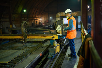 Fototapeta premium Profile view of confident young machine operator wearing filter mask and protective helmet focused on work while standing at spacious production department of modern plant