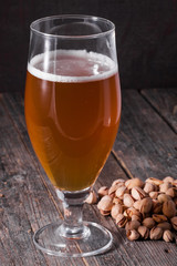 A glass of refreshing light beer and a scattered handful of pistachios on a table of old textured wooden boards.