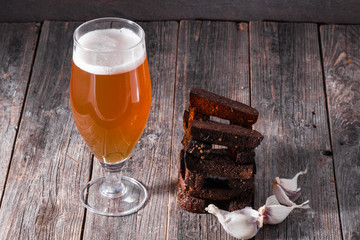A glass of light beer and fried hot aromatic garlic toasts of black bread on a table made of textured old wooden boards