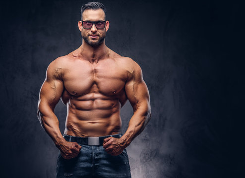 Portrait of a shirtless tall huge male with a muscular body with a stylish haircut and beard, in a sunglasses and jeans, posing in a studio.